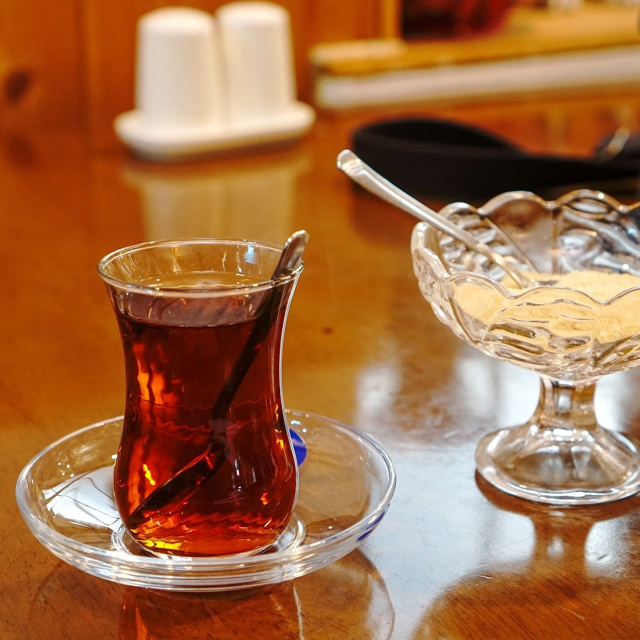 Shapely glass filled with English breakfast tea. A spoonful of sugar may have been stirred into the tea as suggested by spoon and sugar bowl next to it.