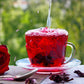 Hibiscus tea overflowing in a glass mug and saucer set. A deep red tea frothing at the top while being poured into the mug. Dried hibiscus flowers decorate the table as does a fresh red rose.