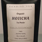 A close-up of a white pouch of Sunrise Tea Organic Hojicha Tea Powder, showing the text 'Hojicha', 'Organic', and 'Tea Powder' in black lettering.