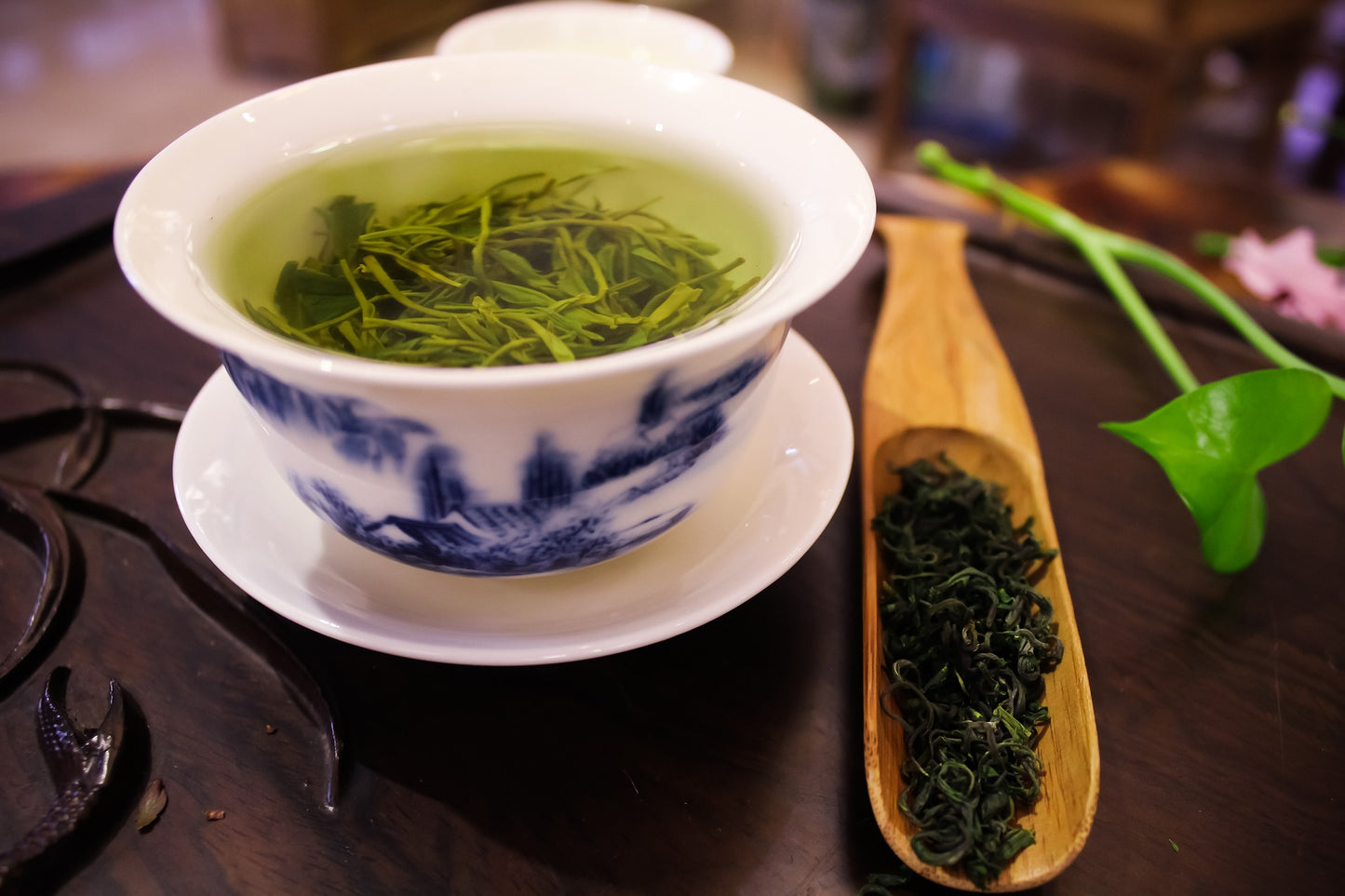 Pretty Chinese tea cup in saucer. Next to it is a large elongated spoon with oval spoon head carved out of wood which contains loose leaf tea. 