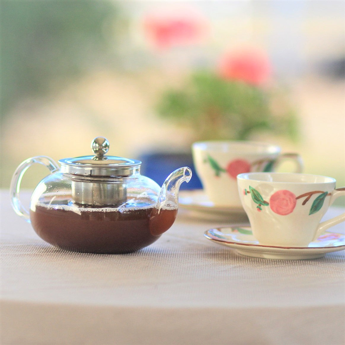 Pretty glass teapot with handle and curved spout filled with black tea ready for serving into accompanying tea cups and saucers with peach design.