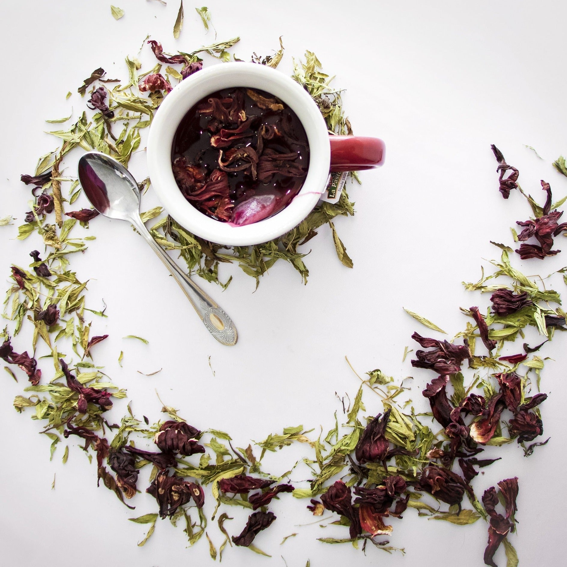 Artistically stylised mug of hibiscus tea from a top down view. Dried hibiscus flowers and leaves strewn in a swirl around a mug of tea. Background is a contrasting white colour.