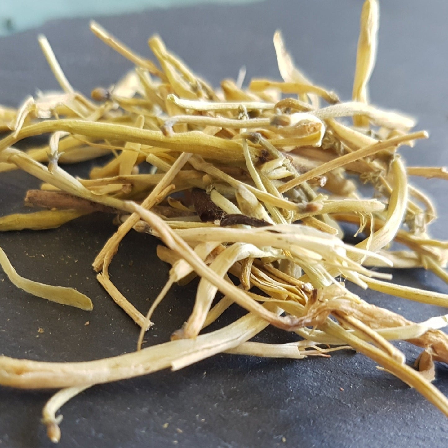 Honeysuckle tea flowers on grey slate tile. Colour is pale yellow. Flowers are dried so long and stringy. Said to eliminate inflammation in the respiratory tracts associated with bronchitis and sore throat. Also helps reduce blood pressure.