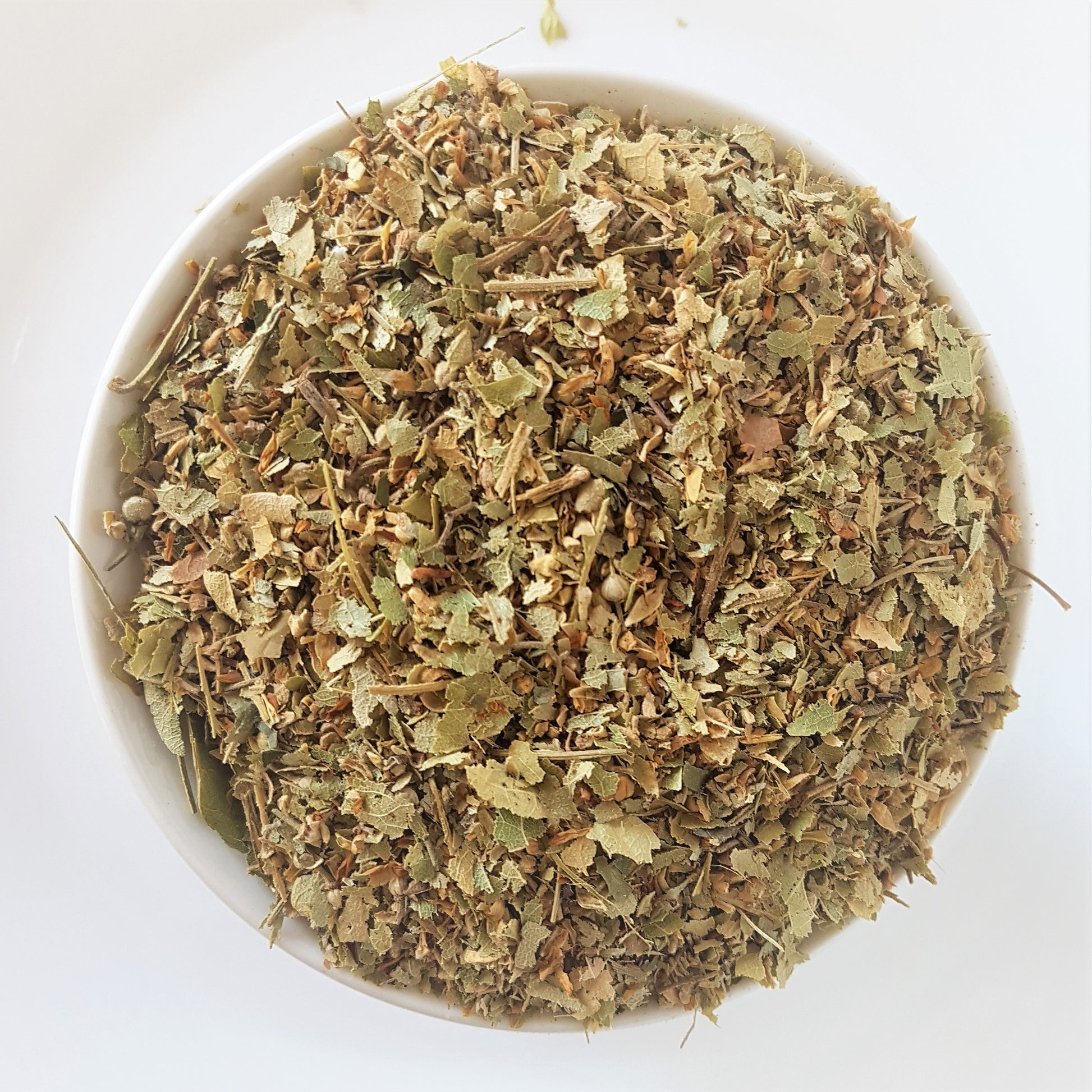 Dried Linden flower tea in a bowl against a white background. Top down view provided. Looks like a mixture of linden flowers and leaves and bits of back of the linden tree. Colours are light green and brown.  Medicinal herbal tea mix which is said to assist against stress, anxiety, inflammation, pain, and high blood pressure. Diuretic effect also.