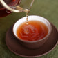 Pu er tea being poured out of a glass tea pot into a brown tea cup  sitting in a matching saucer.  Brewed tea is an attractive orangy brown shade. Tea is rippling in the tea cup as the beverage is being poured out.