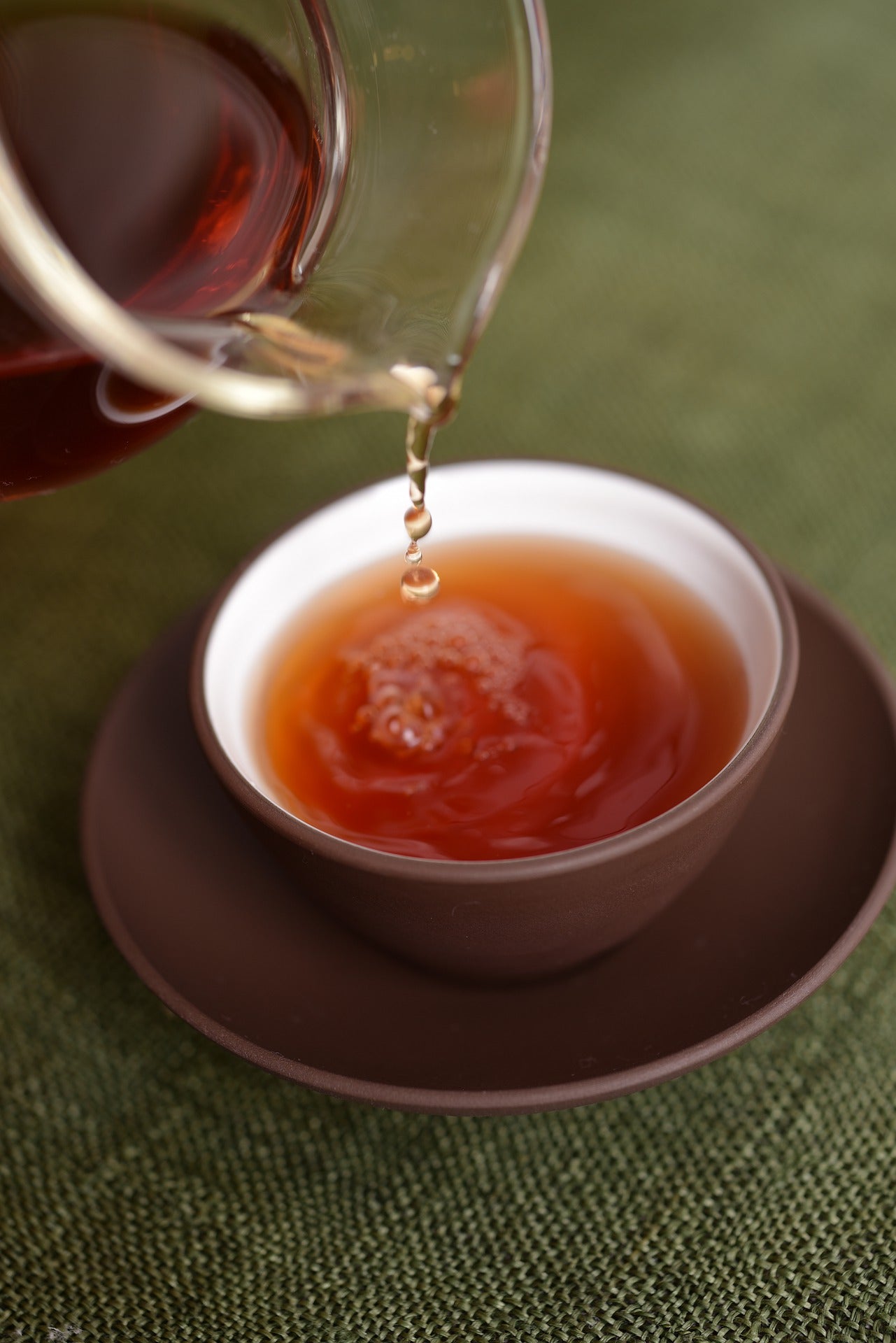 Pu er tea being poured out of a glass tea pot into a brown tea cup  sitting in a matching saucer.  Brewed tea is an attractive orangy brown shade. Tea is rippling in the tea cup as the beverage is being poured out.